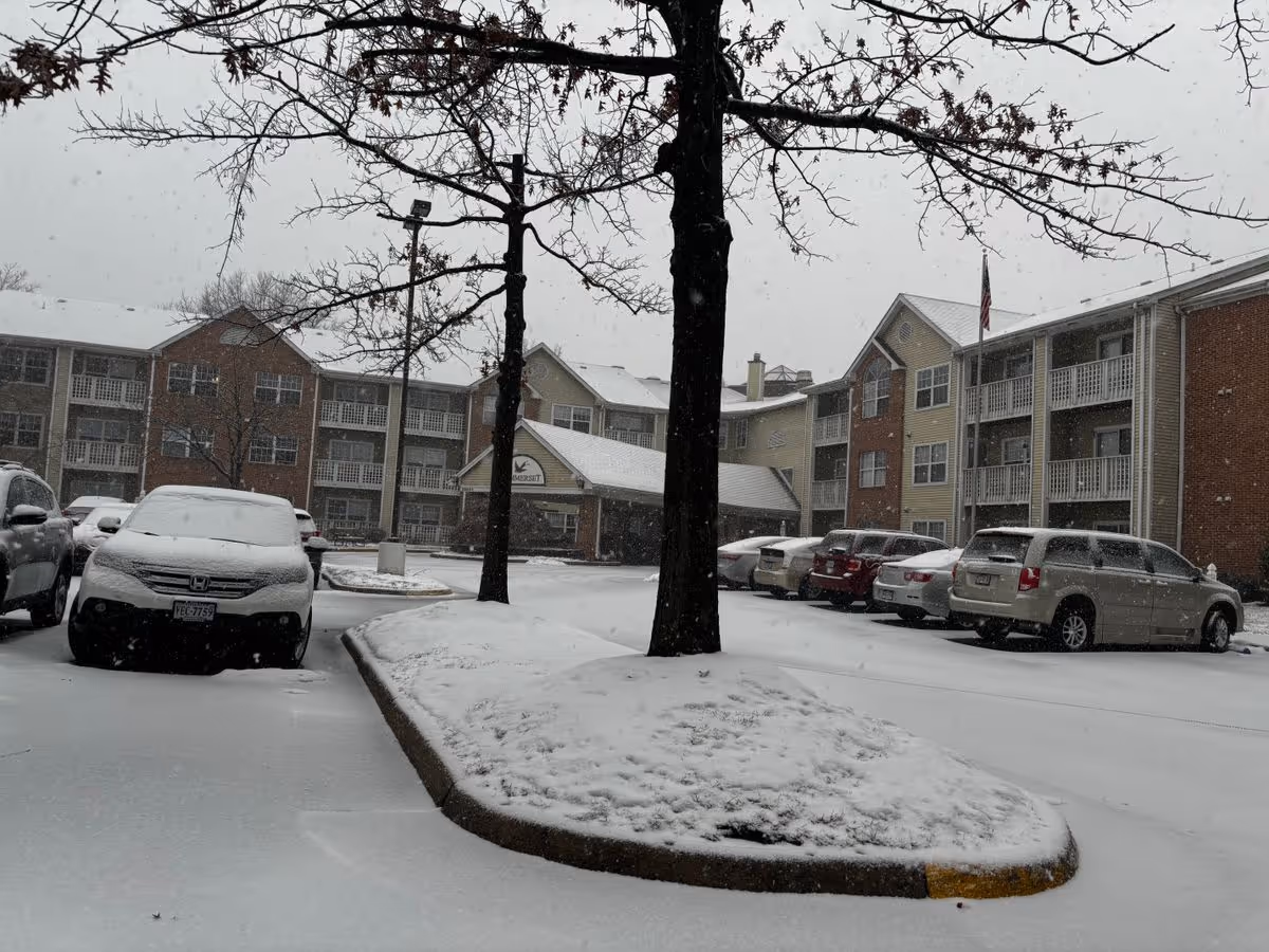 Snow-covered parking lot and the front entrance of Sommerset Retirement Community with cars and bare trees.