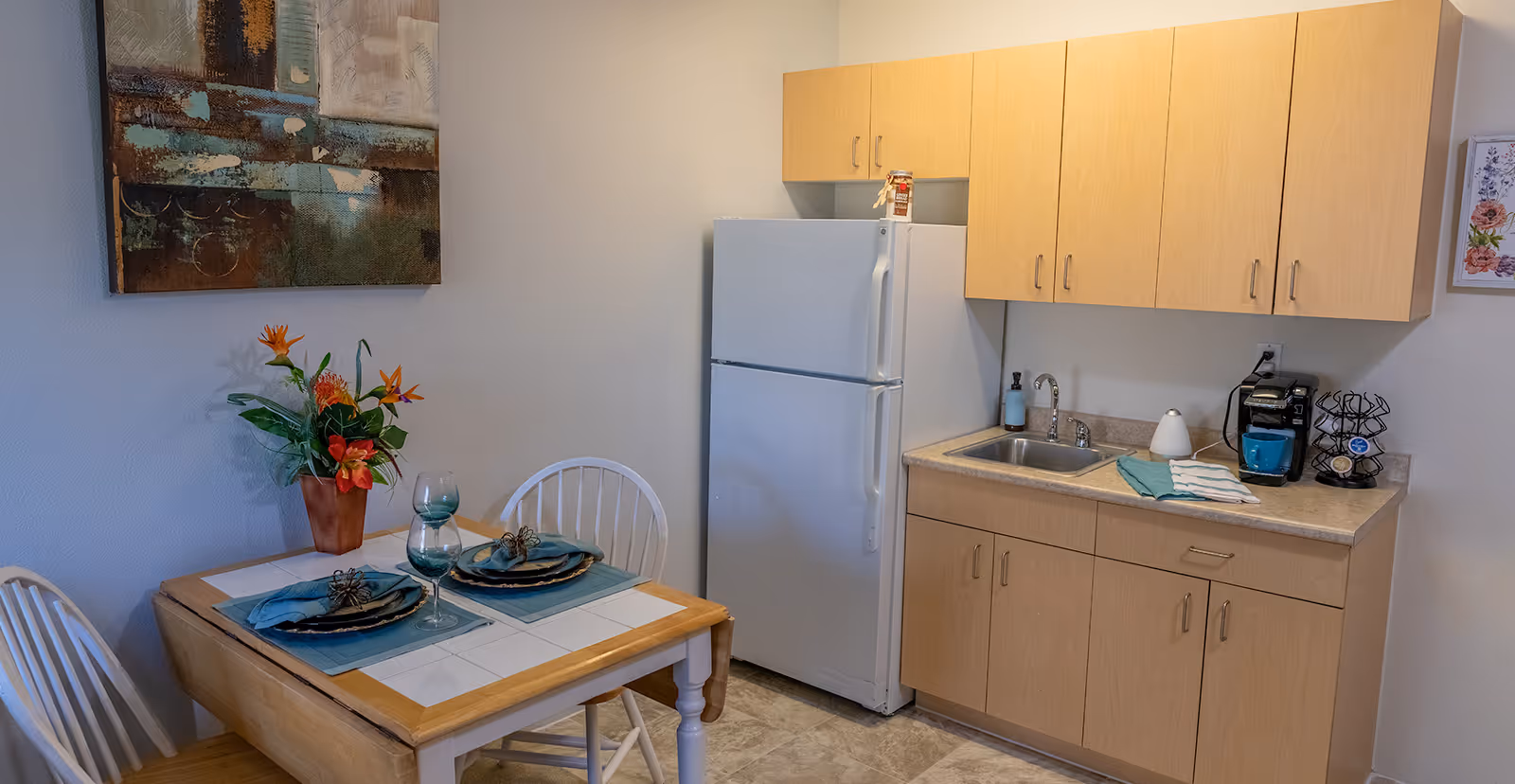 A small kitchen area with light wood cabinets, a white refrigerator, a sink, and a coffee maker on the countertop. Next to the kitchen is a small dining table set for two with blue placemats, plates, napkins, and wine glasses. A vase with colorful flowers and a large abstract painting are on the wall above the table.