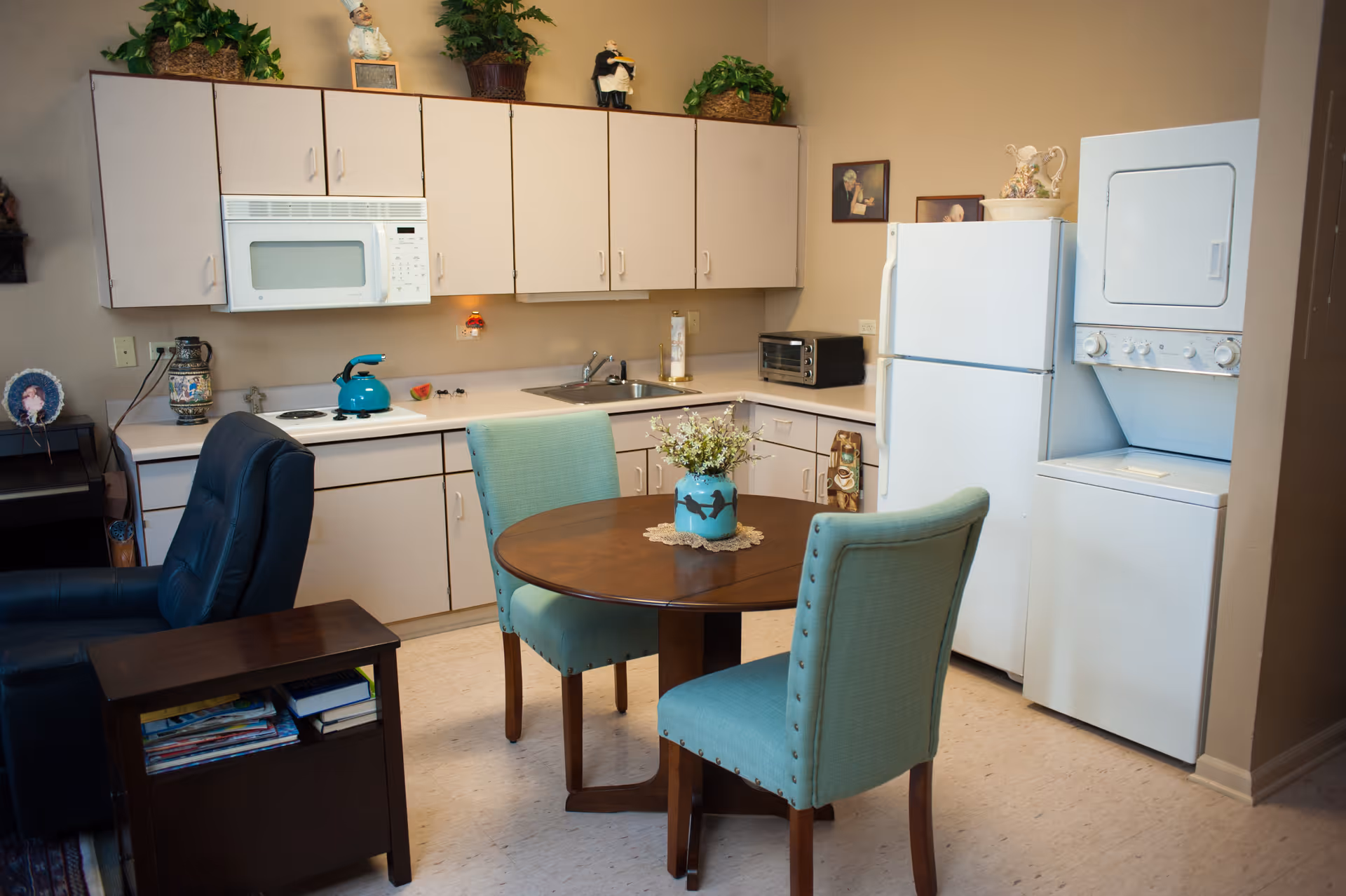 A small kitchen area with beige cabinets, a white microwave, stove, and refrigerator. A stacked washer and dryer unit is next to the refrigerator. In front of the kitchen is a round wooden table with two light blue upholstered chairs. A black recliner chair and a small side table with magazines are also visible. The kitchen counter has a blue kettle, a toaster oven, and some decorative items including plants and figurines.