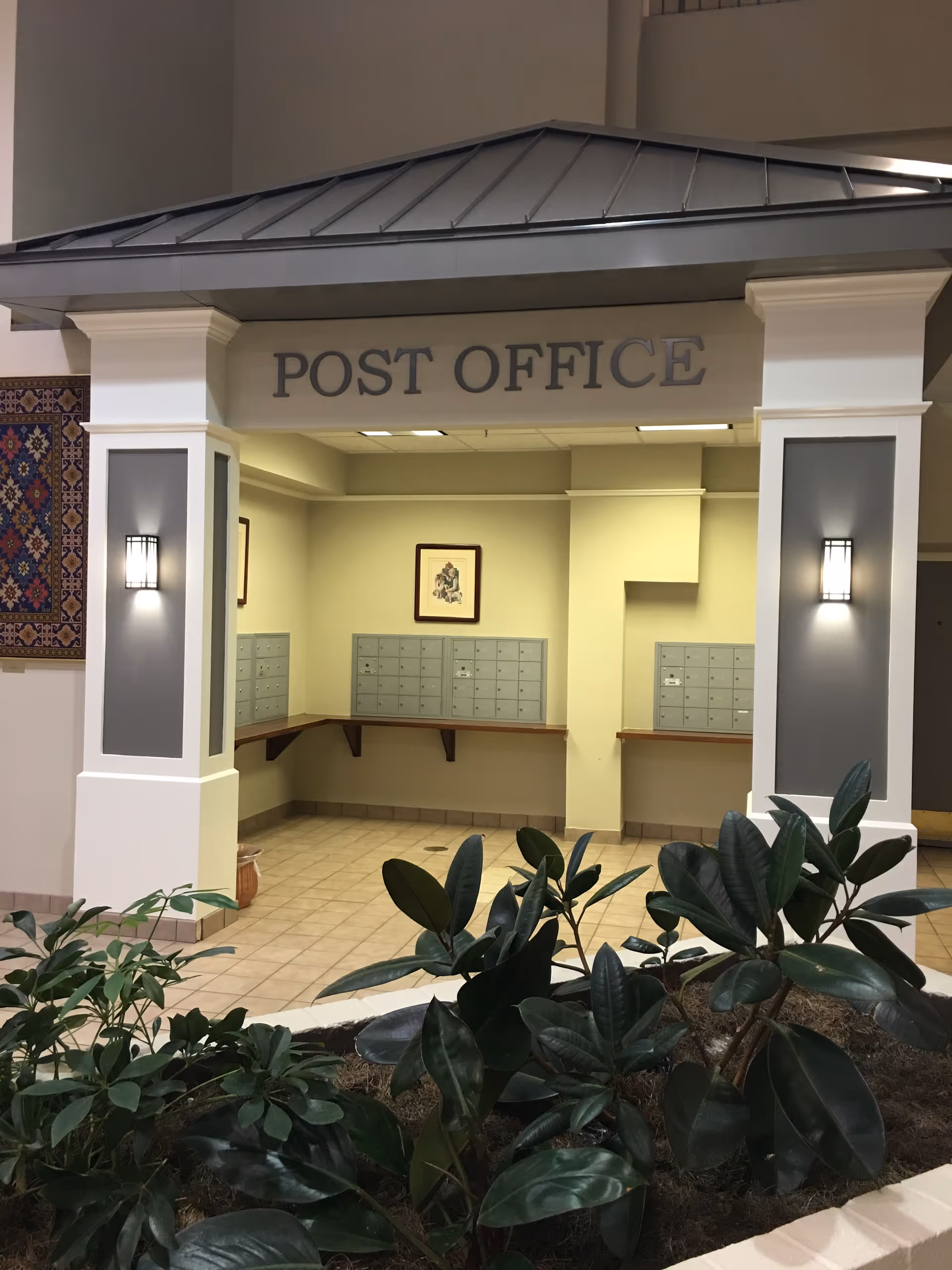 Indoor post office area with multiple mailboxes mounted on the walls under a roofed structure labeled 'POST OFFICE'. There are two wall-mounted lights on white pillars and some green plants in the foreground.