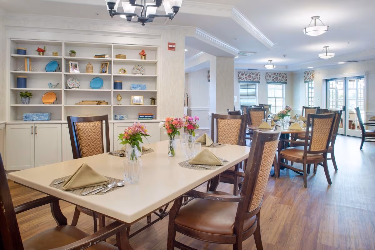 A bright dining room with several tables set with beige napkins, glasses, silverware, and small flower arrangements. The room features wooden chairs with woven backs, wood flooring, and large windows with floral valances allowing natural light to enter. A built-in white shelving unit with decorative plates, books, and small plants is visible on the left wall.