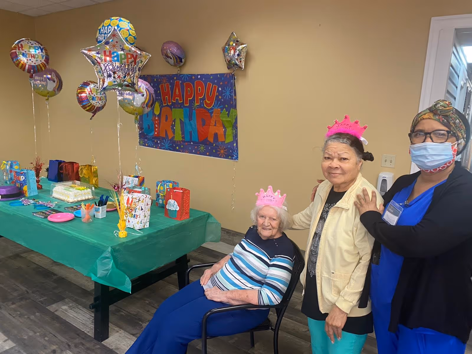 A birthday celebration in a senior living facility room with a table covered in a green tablecloth, decorated with birthday balloons, gift bags, and a birthday cake. Three women are present: one elderly woman sitting in a chair wearing a pink birthday crown, another elderly woman standing beside her also wearing a pink birthday crown, and a staff member wearing a mask and headscarf standing next to them.