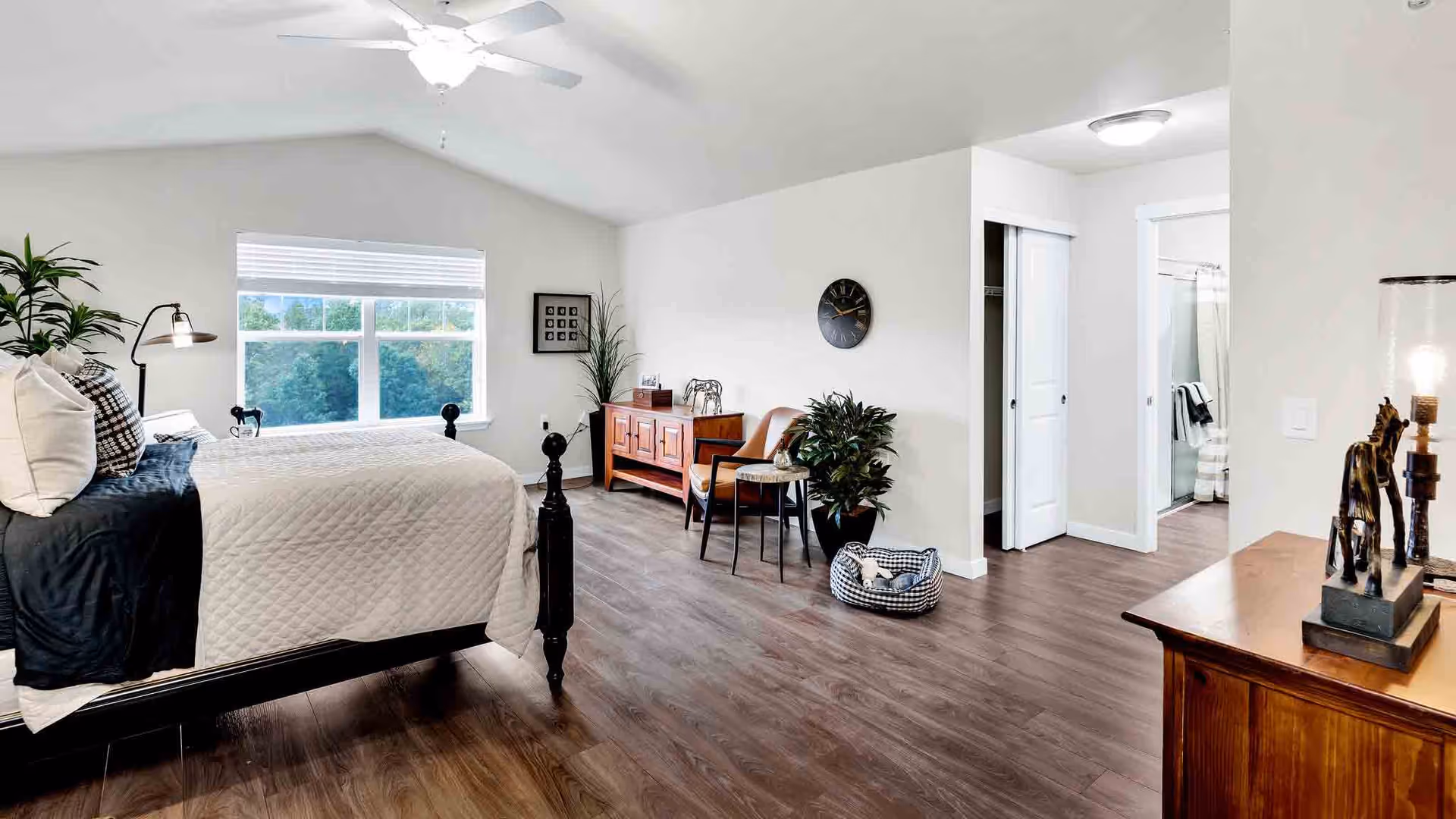 Bright, spacious bedroom with a bed, wooden furniture, plants, and an open doorway to a bathroom.