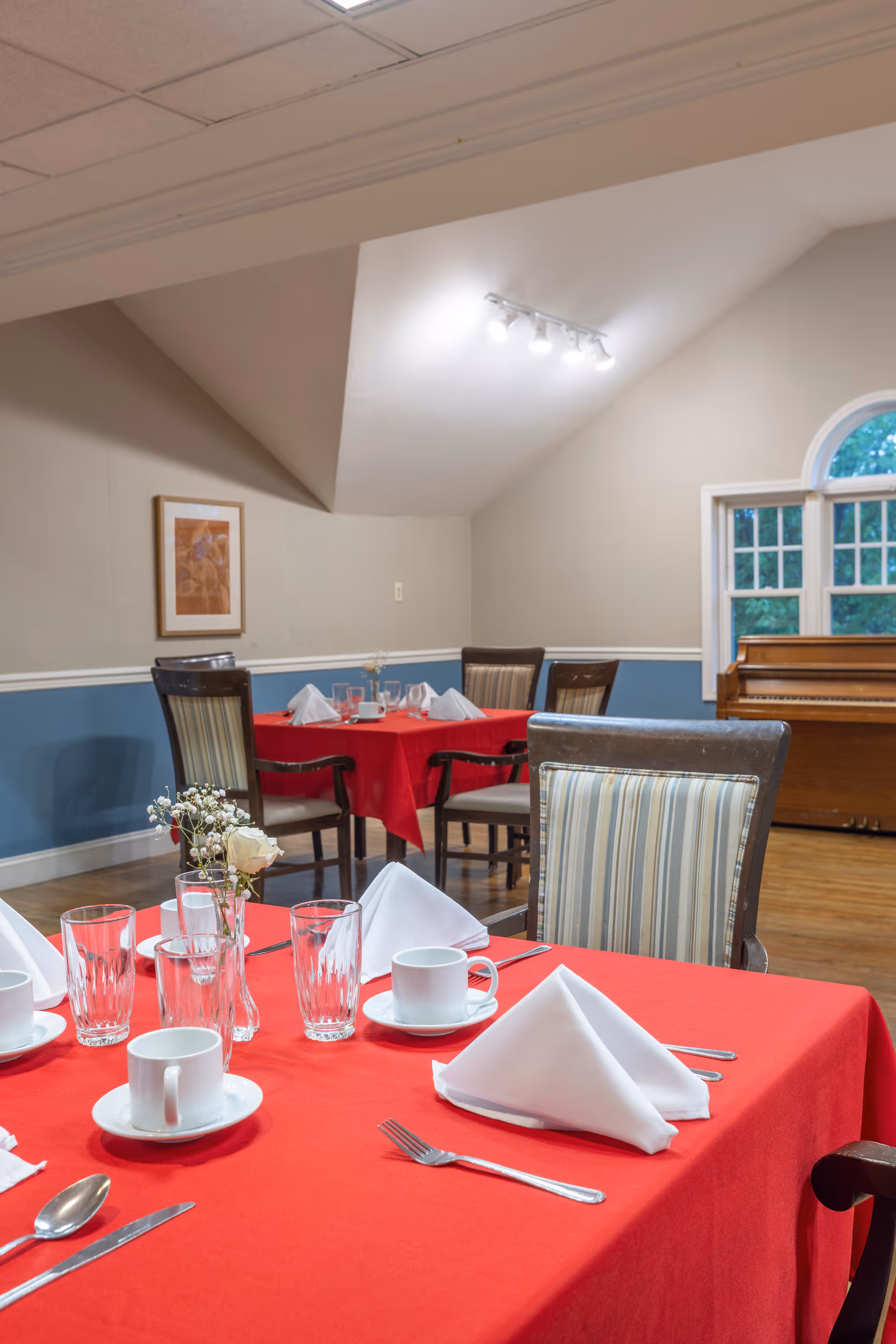 Dining room with tables covered in red tablecloths, set with white napkins, cups, glasses, and silverware. Chairs with striped upholstery surround the tables. A piano is visible near a window in the background.
