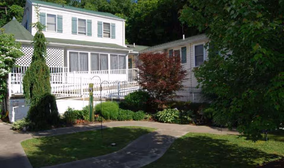 White two-story house with a screened porch, accessibility ramp and a landscaped front yard with trees and shrubs.