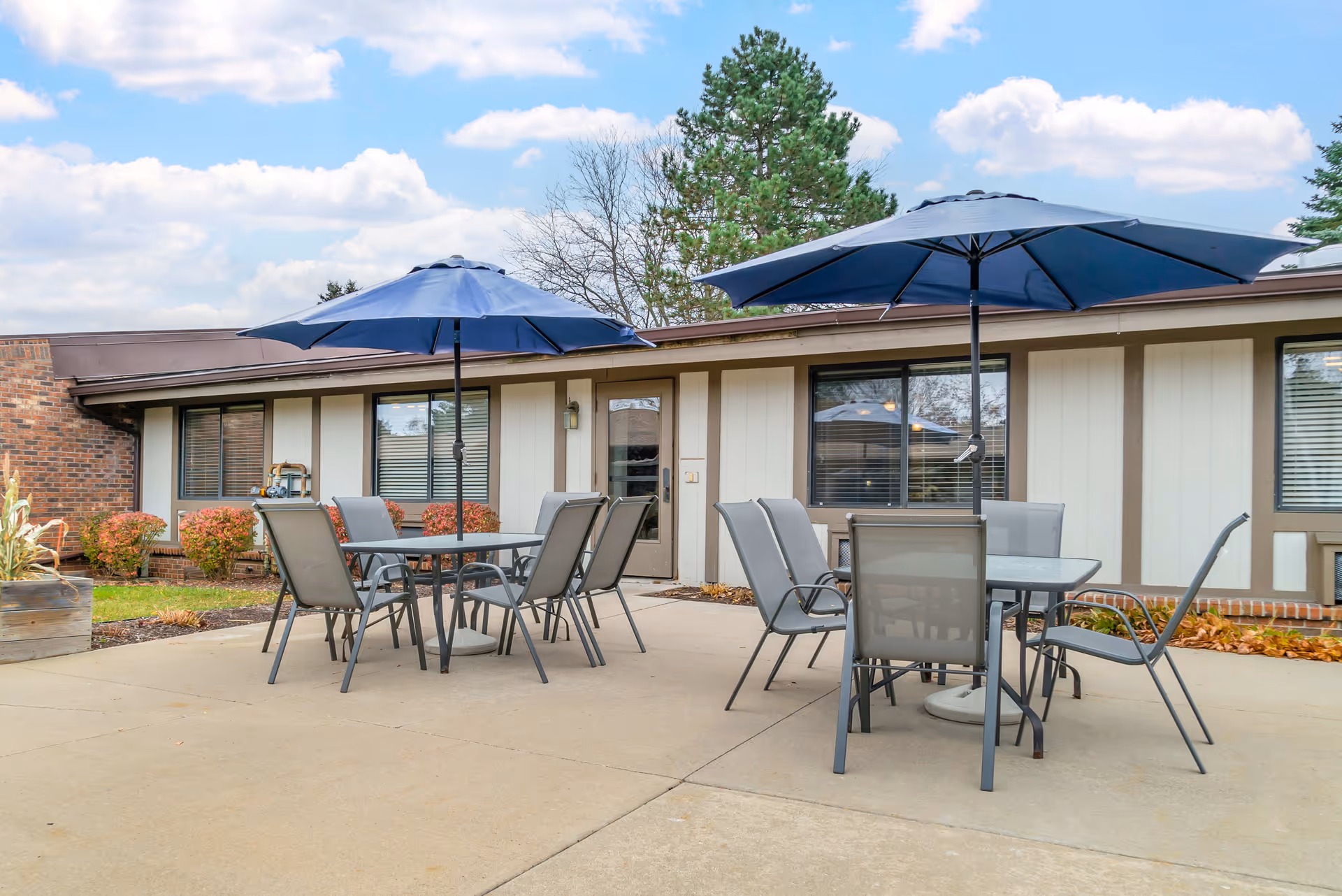 Outdoor patio area at Brookdale Farmington Hills with two tables, each surrounded by six chairs and shaded by large blue umbrellas. The patio is adjacent to a building with large windows and a door, with some landscaping including bushes and trees in the background under a partly cloudy sky.