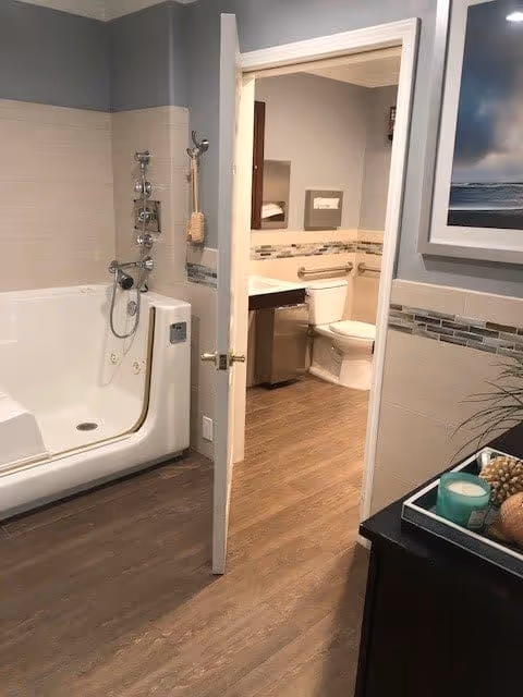 View of a bathroom with a walk-in bathtub on the left and an open door leading to a toilet and sink area. The bathroom has wood-style flooring, light-colored tiled walls with decorative trim, and a framed picture on the wall. A small table with decorative items is visible on the right side.