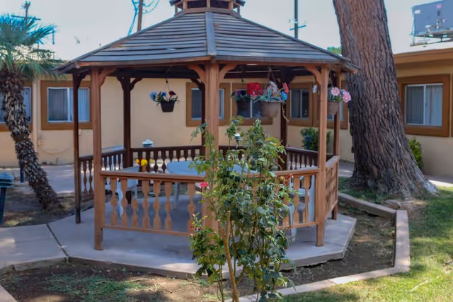 A wooden gazebo with a hexagonal roof and railing situated in an outdoor courtyard area with a tree, plants, and a building with windows in the background.