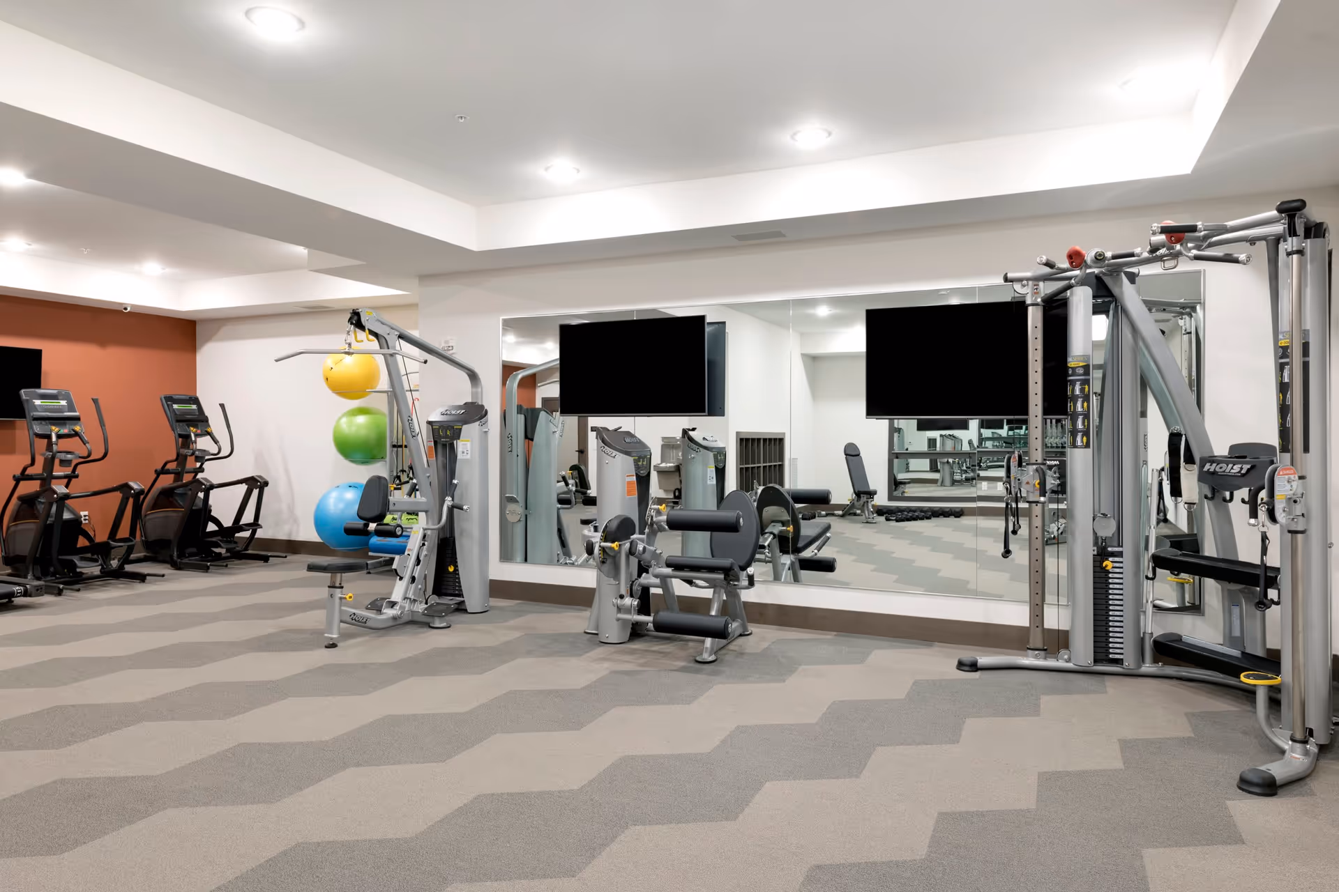 A modern fitness room with exercise equipment including treadmills, weight machines, and colorful exercise balls. The room has a patterned carpet, large mirrors on the wall, and two flat-screen TVs mounted above the equipment.