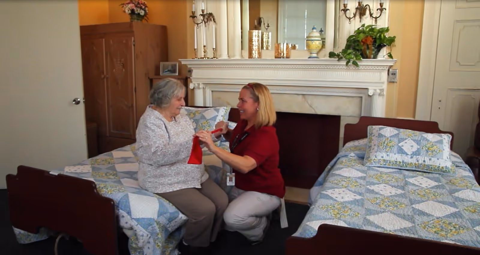 A caregiver kneels between two quilt-covered twin beds helping an elderly resident in a cozy room with a fireplace mantel and plants.
