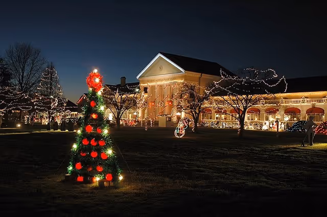 A nighttime outdoor scene of a large building decorated with Christmas lights. In the foreground, there is a Christmas tree adorned with red and green lights. The building and surrounding trees are also decorated with white string lights, creating a festive holiday atmosphere.