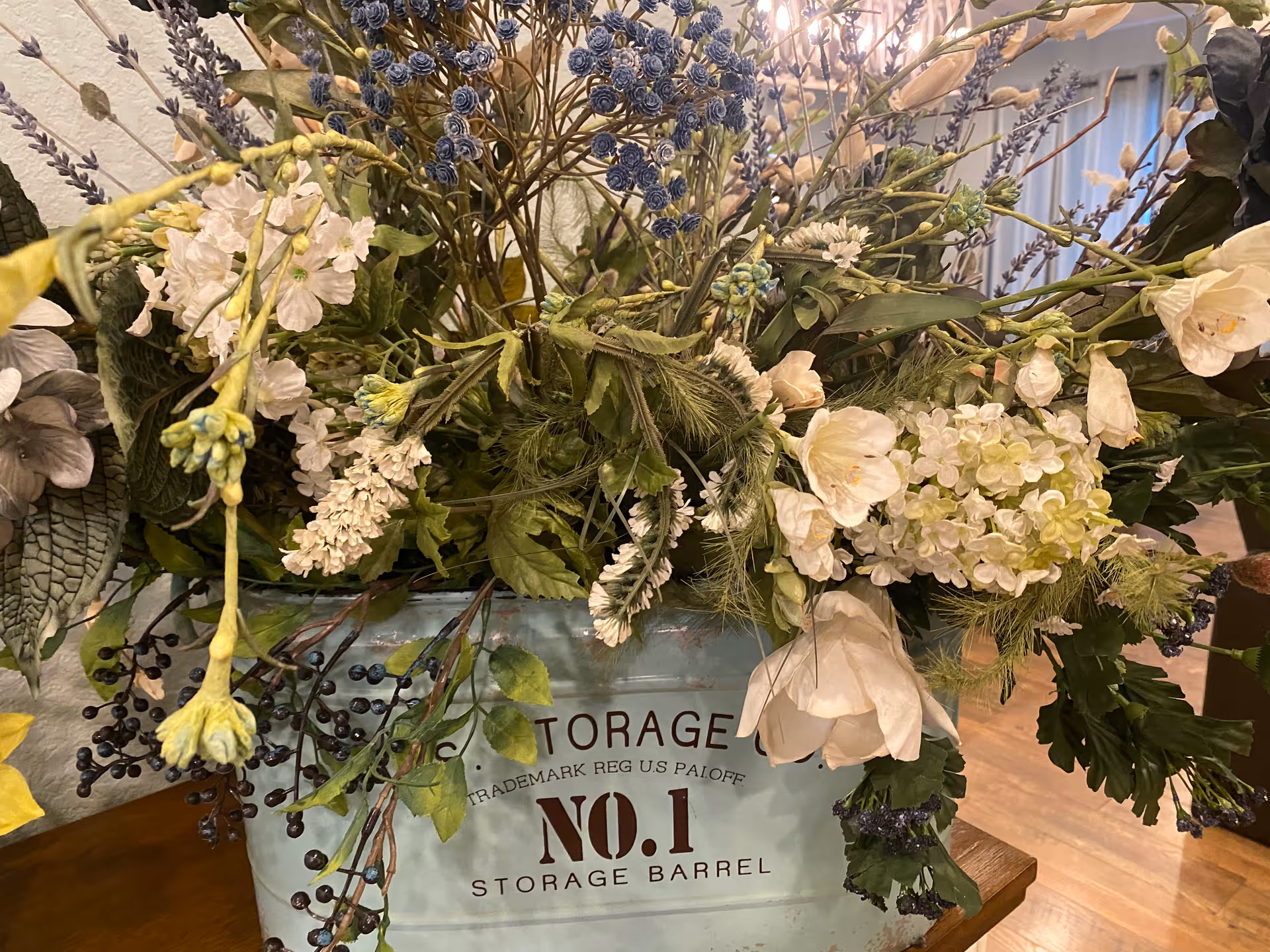 A large arrangement of white and purple flowers and greenery in a blue metal container labeled 'NO.1 STORAGE BARREL' sitting on a wooden surface.