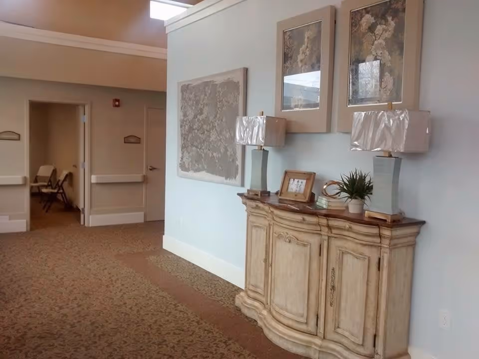 Interior hallway of a senior living facility with beige patterned carpet and light blue walls. A wooden sideboard with two lamps, a small plant, books, and a framed photo is against the wall. Above the sideboard are three framed floral artworks. Two open doorways lead to rooms with chairs visible inside.