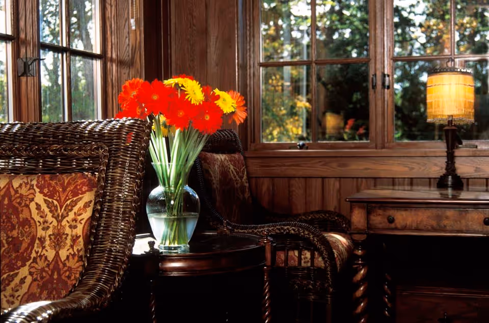 A cozy interior corner with two wicker chairs featuring patterned cushions, a small round wooden table holding a glass vase with bright orange and yellow flowers, and a wooden desk with a vintage-style lamp. Large windows with wooden frames show a view of trees outside.