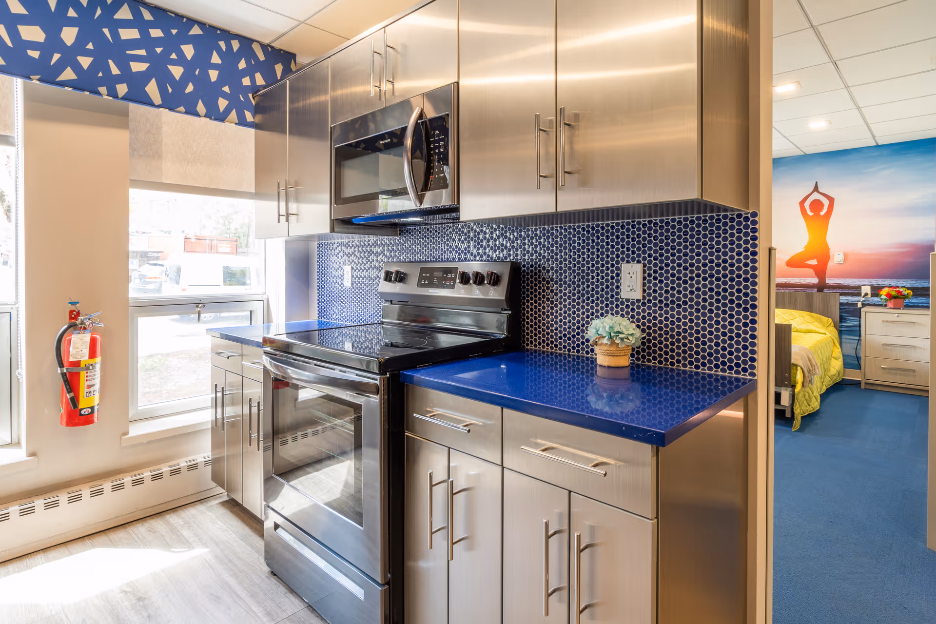 Modern kitchen area with stainless steel cabinets, a black electric stove, and a microwave. The countertop is blue with a matching blue hexagonal tile backsplash. A small potted plant is on the counter. To the left, there are two windows with a fire extinguisher mounted on the wall. In the background, a bedroom with a bed, nightstand, and a wall mural of a person doing yoga at sunset is visible.