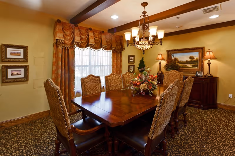 A formal dining room with a large polished wooden table surrounded by eight upholstered chairs. The room features patterned carpet, ornate curtains with valances over a window, a chandelier hanging above the table, framed artwork on the walls, and a sideboard with two table lamps and a landscape painting.