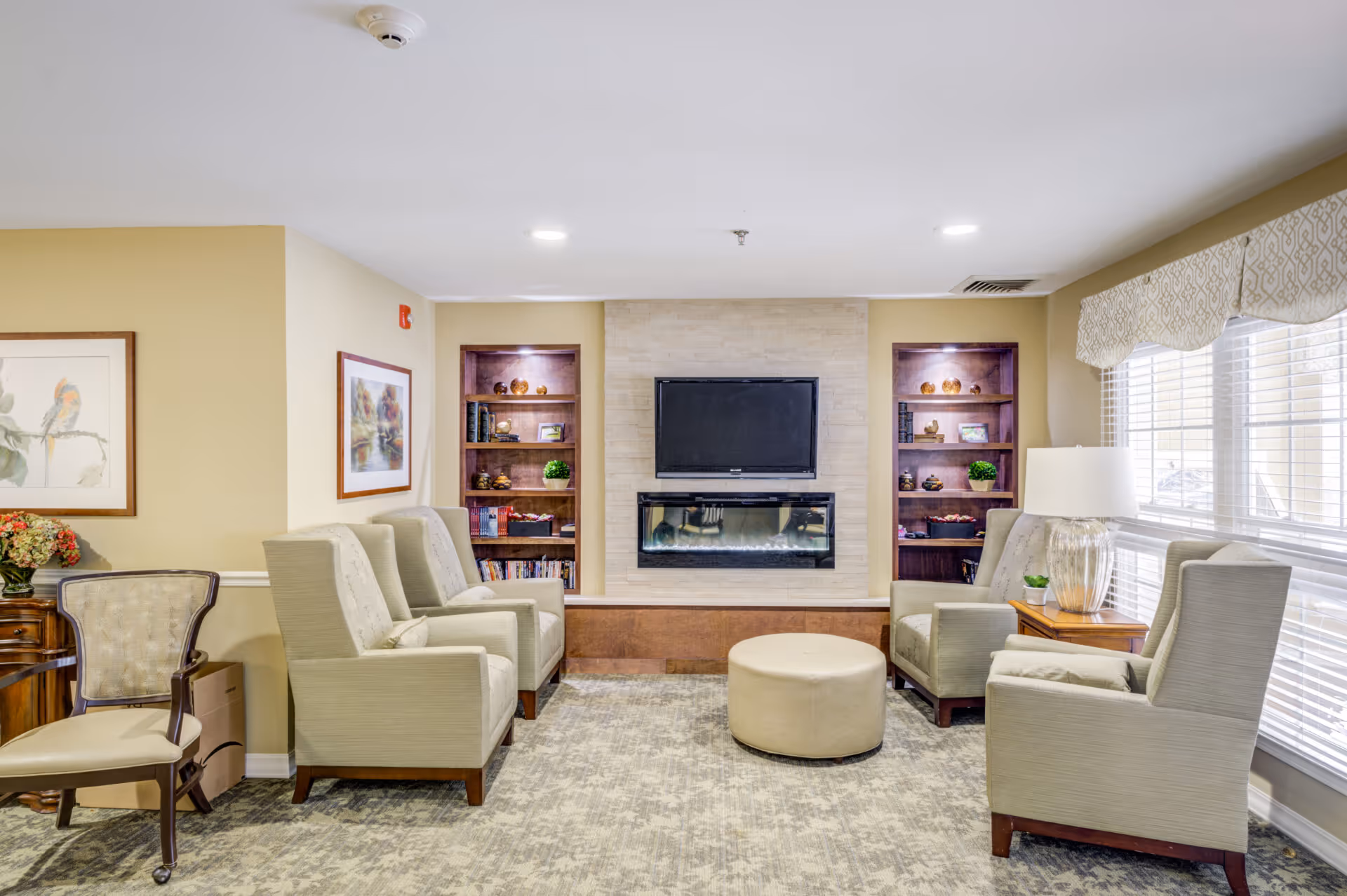 A cozy senior living room with four beige armchairs arranged around a round beige ottoman. The room features a wall-mounted flat screen TV above a modern electric fireplace, flanked by built-in wooden shelves with decorative items and books. Large windows with patterned valances allow natural light to fill the space. There is a side table with a lamp and a small plant next to one of the armchairs, and framed artwork on the walls.