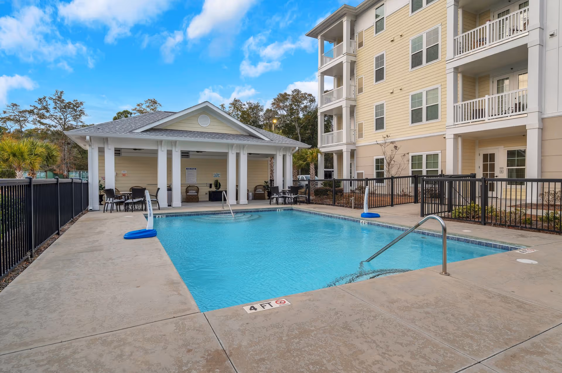 Outdoor swimming pool area at a senior living facility with a covered seating area featuring tables and chairs. The pool is surrounded by a concrete deck and black metal fencing. A multi-story beige building with balconies is visible in the background under a partly cloudy blue sky.
