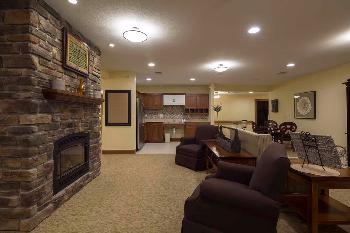 Interior view of a senior living facility common area featuring a stone fireplace on the left, two dark armchairs, a beige sofa, wooden side tables, and a kitchen area with wooden cabinets and stainless steel refrigerator in the background. The room is warmly lit with ceiling lights and has beige walls and carpeted flooring.