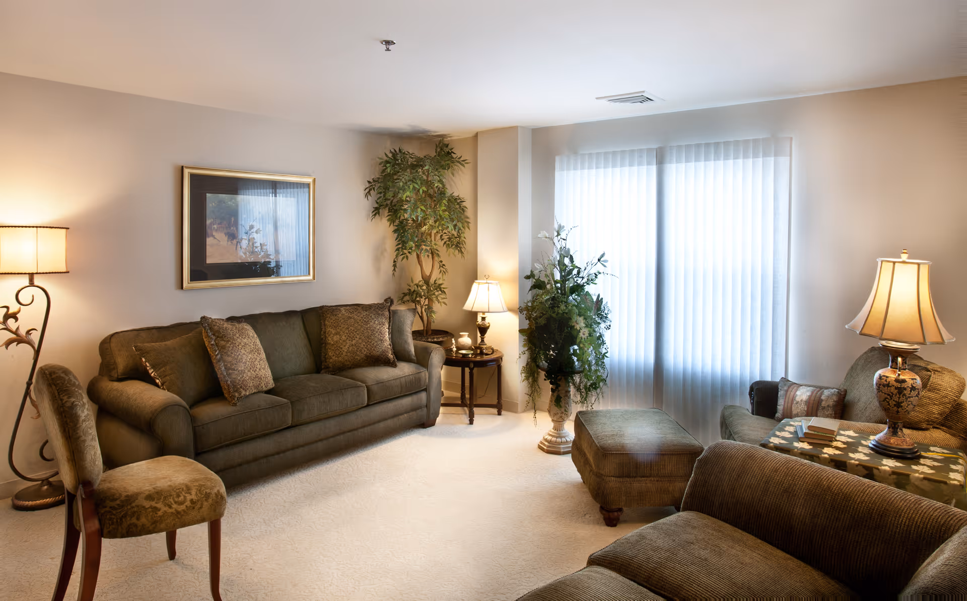 A cozy living room with a green sofa, matching armchairs, and an ottoman. The room is softly lit by two table lamps and a floor lamp. There is a framed picture on the wall, a potted plant in the corner, and a large window with vertical blinds letting in natural light.