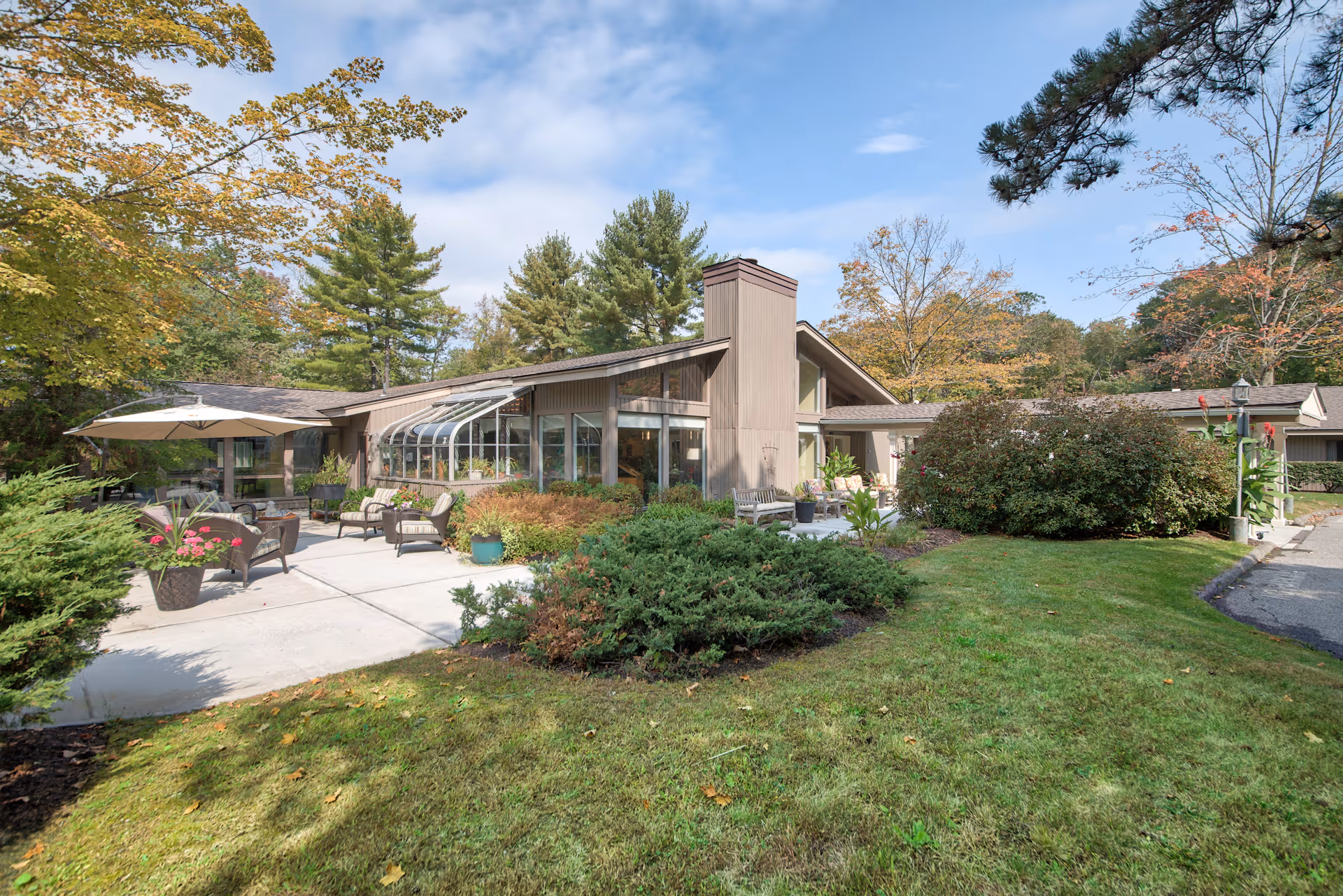 A single-story building with large windows and a glass sunroom, surrounded by green shrubs and trees with autumn foliage. There is a concrete patio area with outdoor seating including chairs, tables, and an umbrella. The sky is partly cloudy.