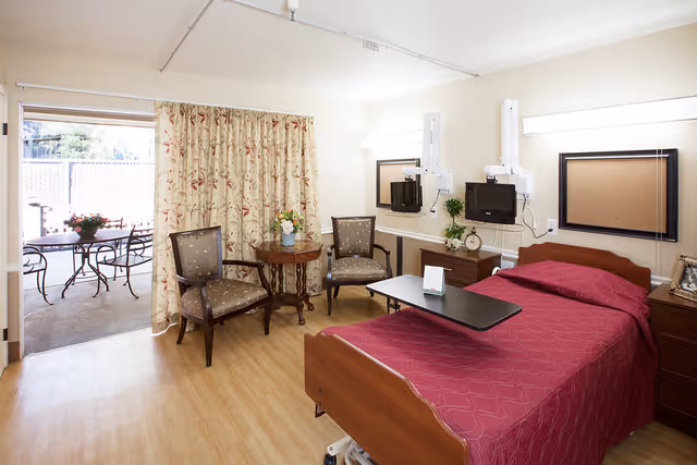 A senior care facility bedroom with a single bed covered in a red quilt, a movable overbed table, two upholstered chairs, a small round wooden table with a flower vase, and a sliding glass door leading to an outdoor patio with a table and chairs. The room has light-colored walls and wood flooring, with two mounted televisions and framed cork boards on the wall.
