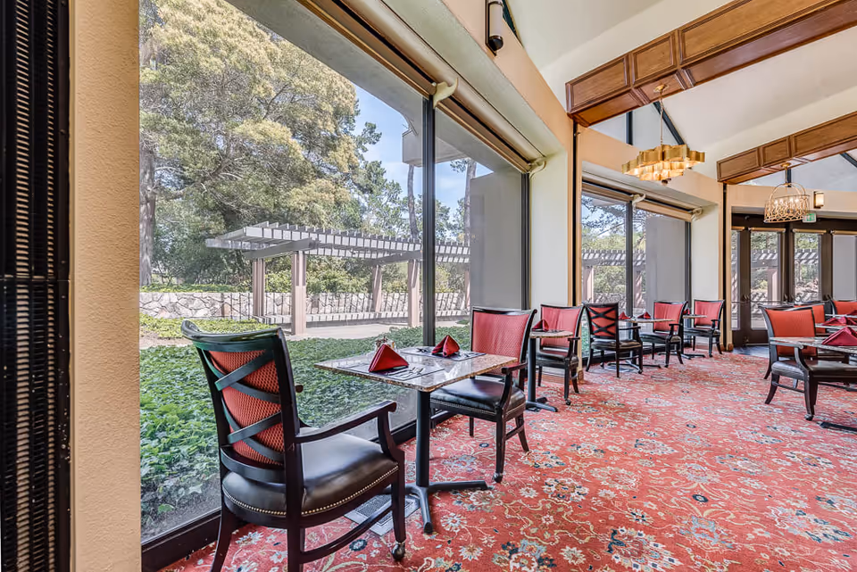 Interior view of a dining area with multiple tables and chairs arranged along large windows overlooking a garden with trees and a pergola. The room features a red patterned carpet, high ceilings with wooden beams, and modern hanging light fixtures.