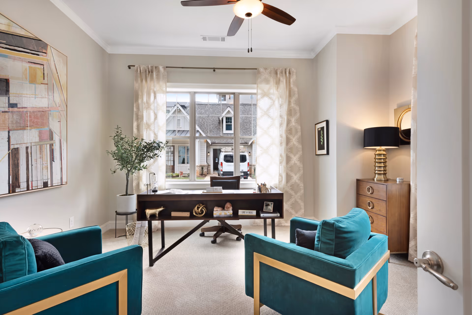 Furnished home office with teal armchairs, a desk facing a large window, and decorative accents.