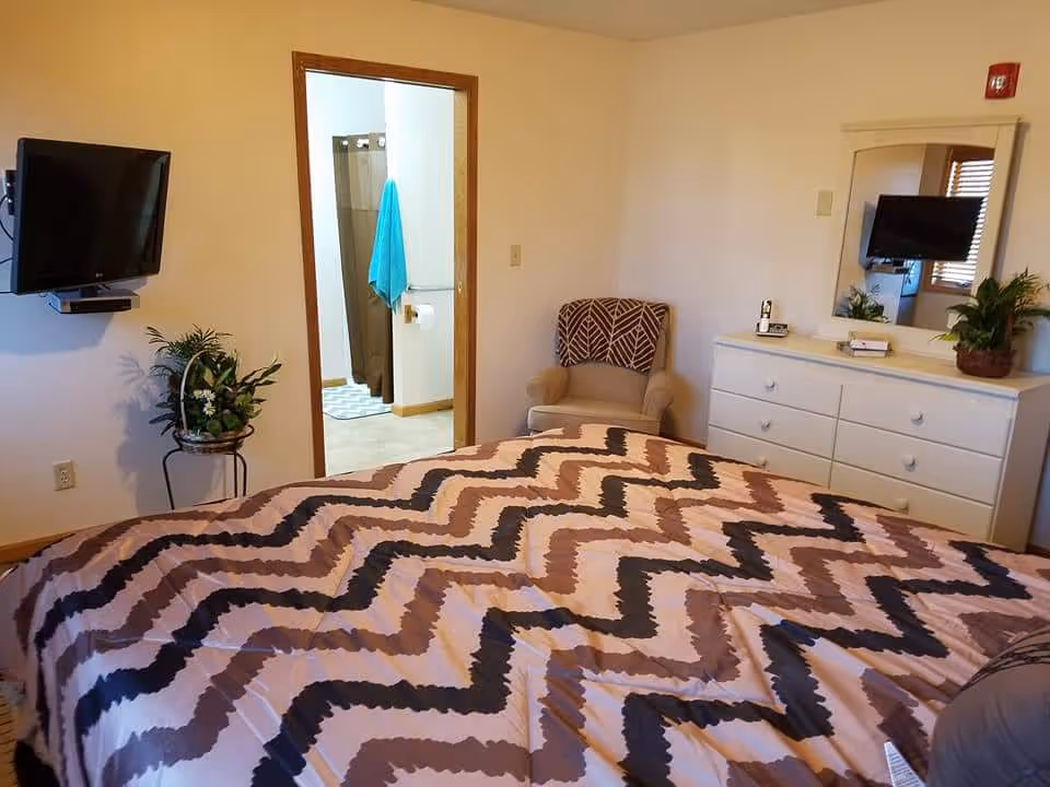A bedroom with a bed covered in a zigzag patterned blanket in shades of brown, beige, and black. There is a beige armchair with a patterned cushion in the corner, a white dresser with a mirror above it, and a mounted flat-screen TV on the wall. A doorway leads to a bathroom where a turquoise towel hangs on a rack.