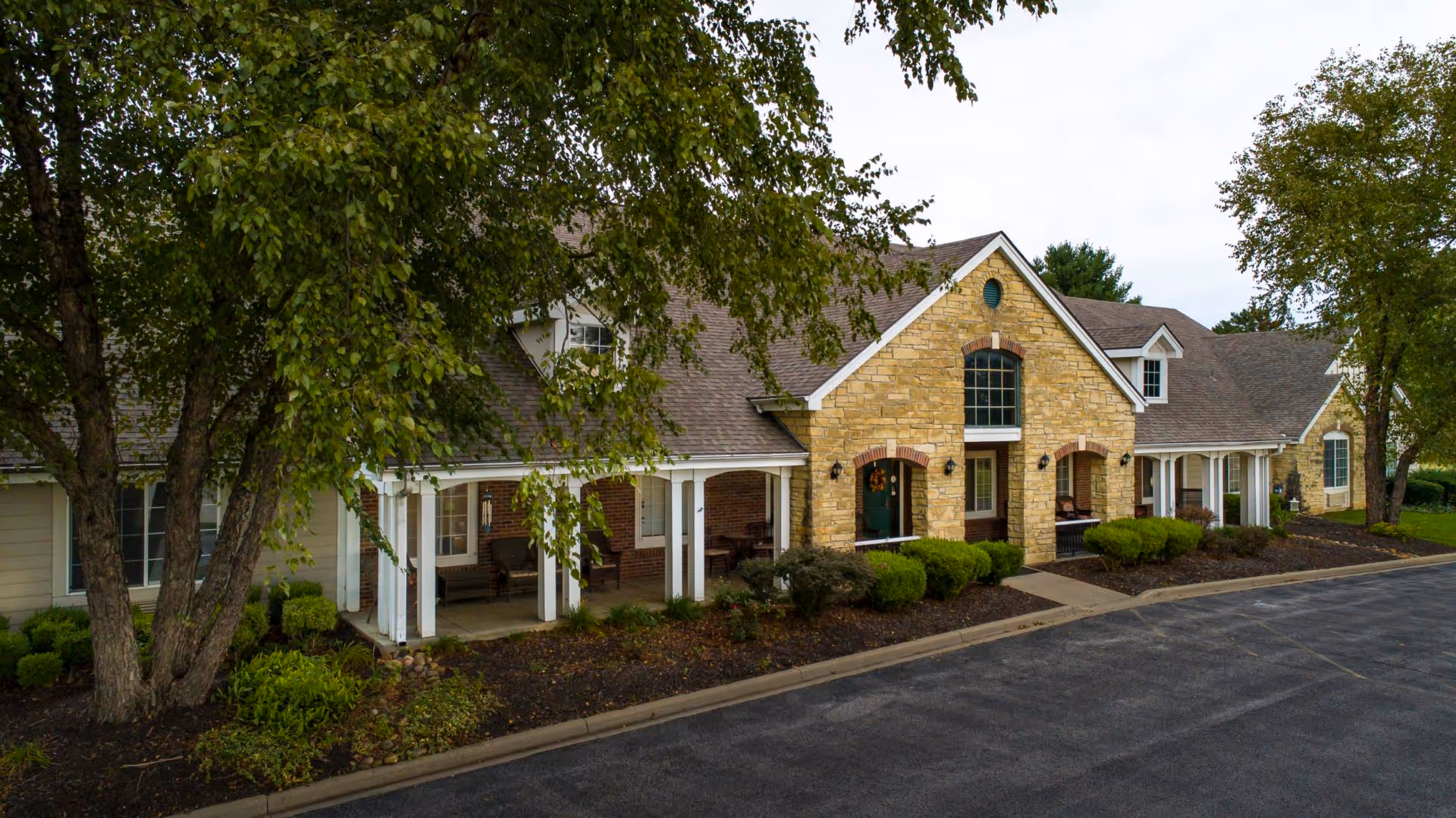 Front exterior of a stone-faced assisted living building with covered porches, landscaping, and a driveway.