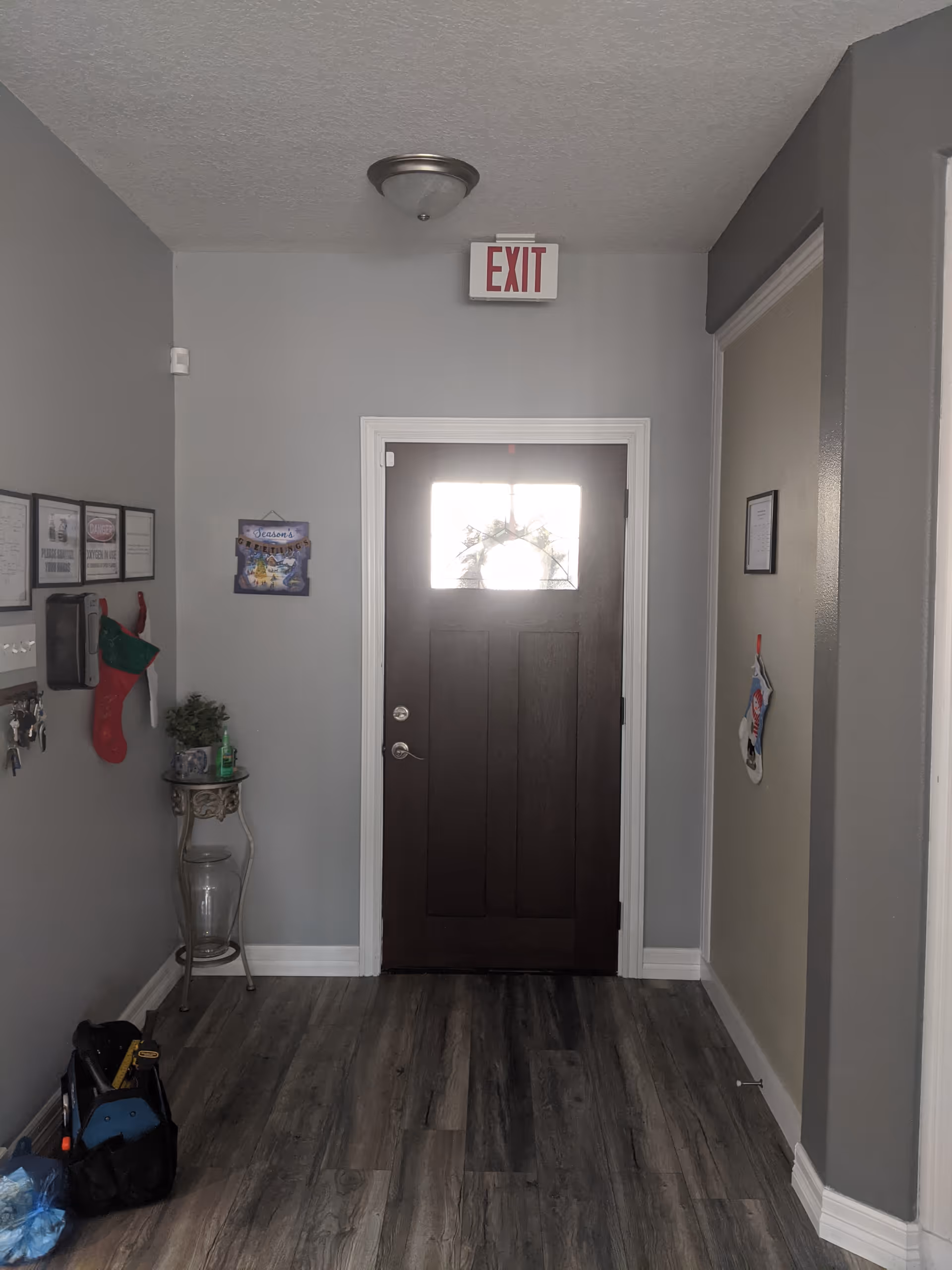Interior view of a hallway with a dark wooden door featuring a small window with a wreath. Above the door is an illuminated exit sign. The walls are painted gray, and the floor has wood-like vinyl planks. On the left wall, there are framed signs, a red Christmas stocking, a small table with a plant and hand sanitizer, and a set of keys hanging. On the right wall, there is a single framed item and another Christmas stocking hanging.