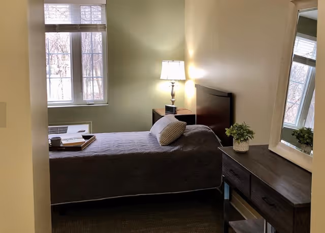 A small, neatly arranged bedroom with a single bed covered in a gray quilt. There is a pillow on the bed and a tray with a cup and a book. A bedside table with a lamp and a clock is next to the bed. A dresser with a potted plant and a large mirror is on the right side of the room. A window with blinds lets in natural light.