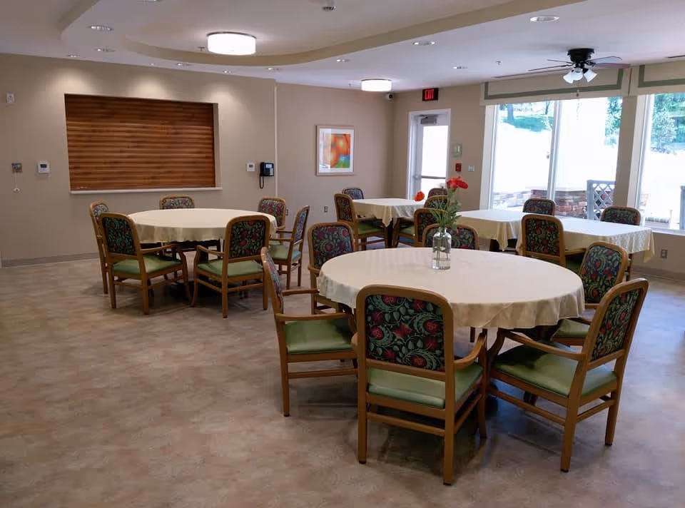 A dining room with several round and rectangular tables covered with white tablecloths. Each table is surrounded by wooden chairs with green cushions and floral-patterned backs. A vase with red flowers is placed on the nearest round table. The room has beige walls, a large window letting in natural light, and ceiling lights.