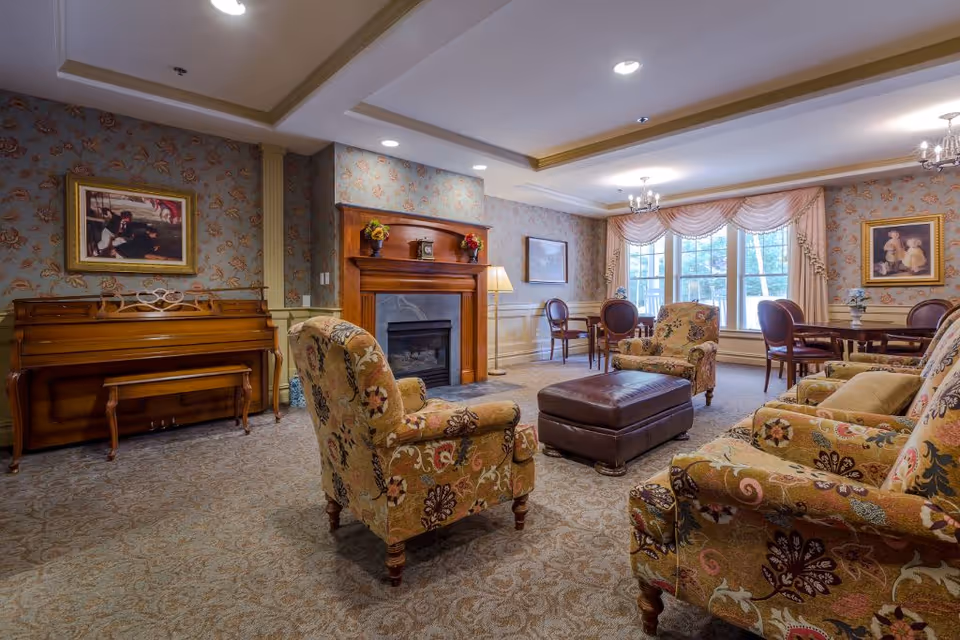 A cozy living room in an assisted living facility featuring floral upholstered armchairs and a matching sofa arranged around a brown leather ottoman. The room has a wooden piano against one wall, a fireplace with a wooden mantel decorated with flowers and a clock, and large windows with elegant drapes letting in natural light. The walls are adorned with framed paintings and the carpet has a subtle patterned design.