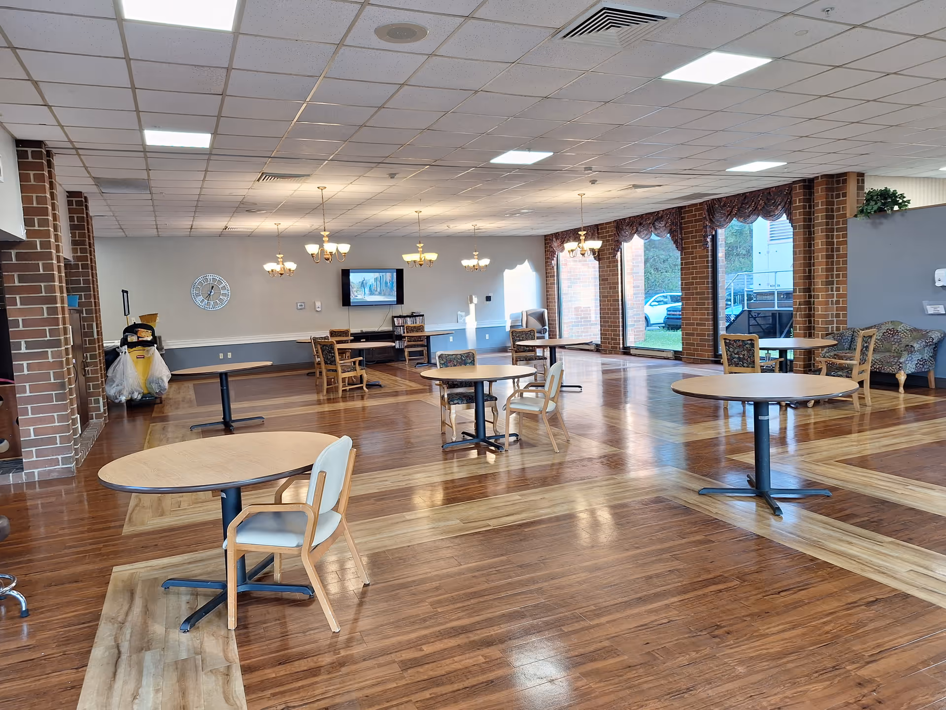 Spacious senior living communal dining/activity room with round tables and chairs, chandeliers, a TV, and large windows with brick columns.