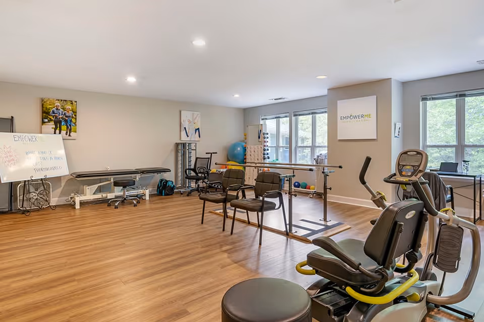 A spacious rehabilitation or physical therapy room with wooden flooring, exercise equipment including a recumbent bike, parallel bars, chairs, therapy balls, and a treatment table. The room has large windows letting in natural light and motivational posters on the walls.