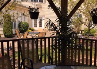View from a covered porch or patio area with a round glass table and chairs in the foreground, overlooking an outdoor garden space with a table and chairs, hanging flower pots, bushes, and trees near a light-colored building.
