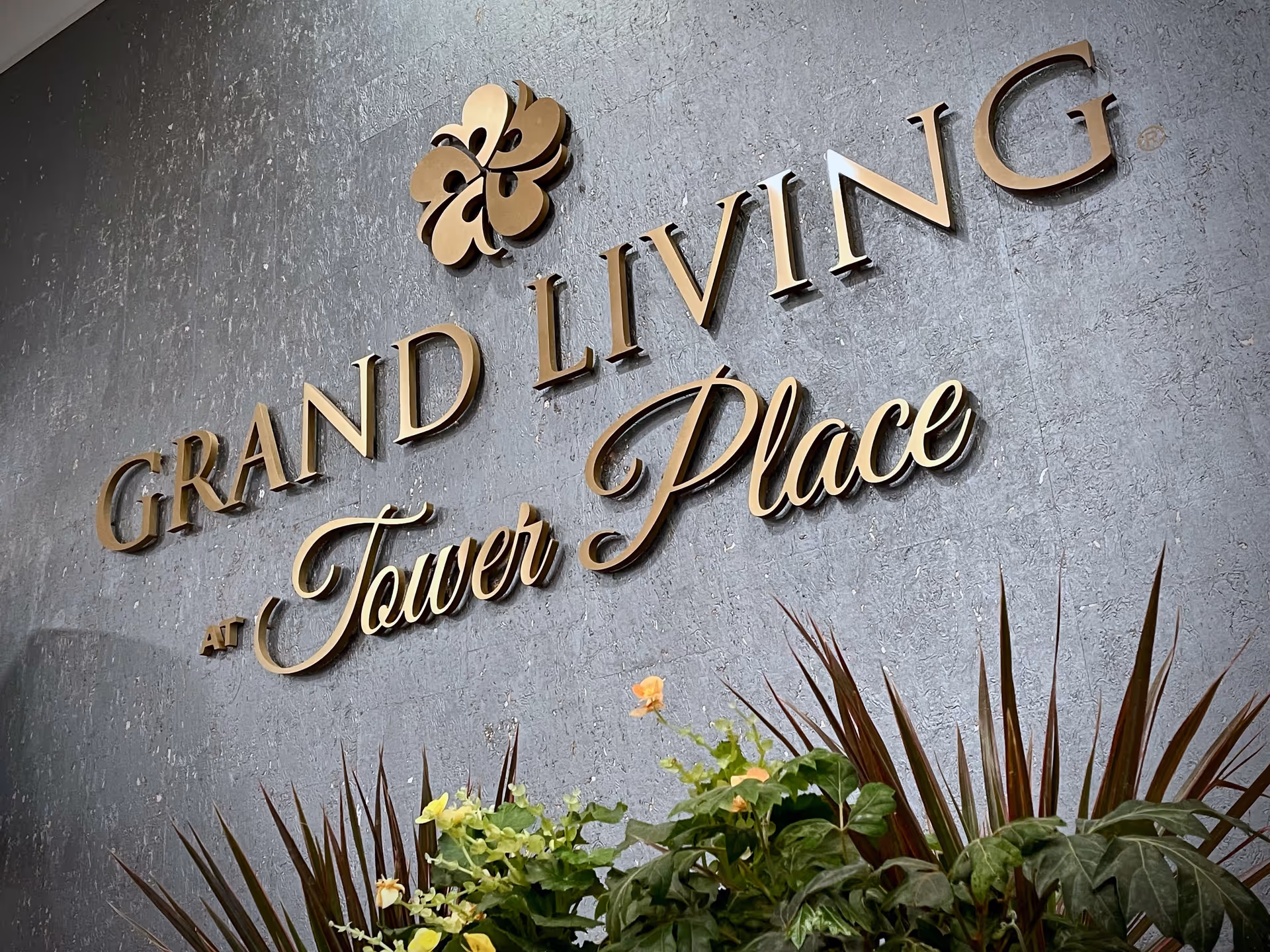 A textured gray wall with gold-colored 3D letters spelling 'GRAND LIVING at Tower Place' accompanied by a gold floral emblem above the text. Below the text, there are green plants with some yellow and orange flowers.