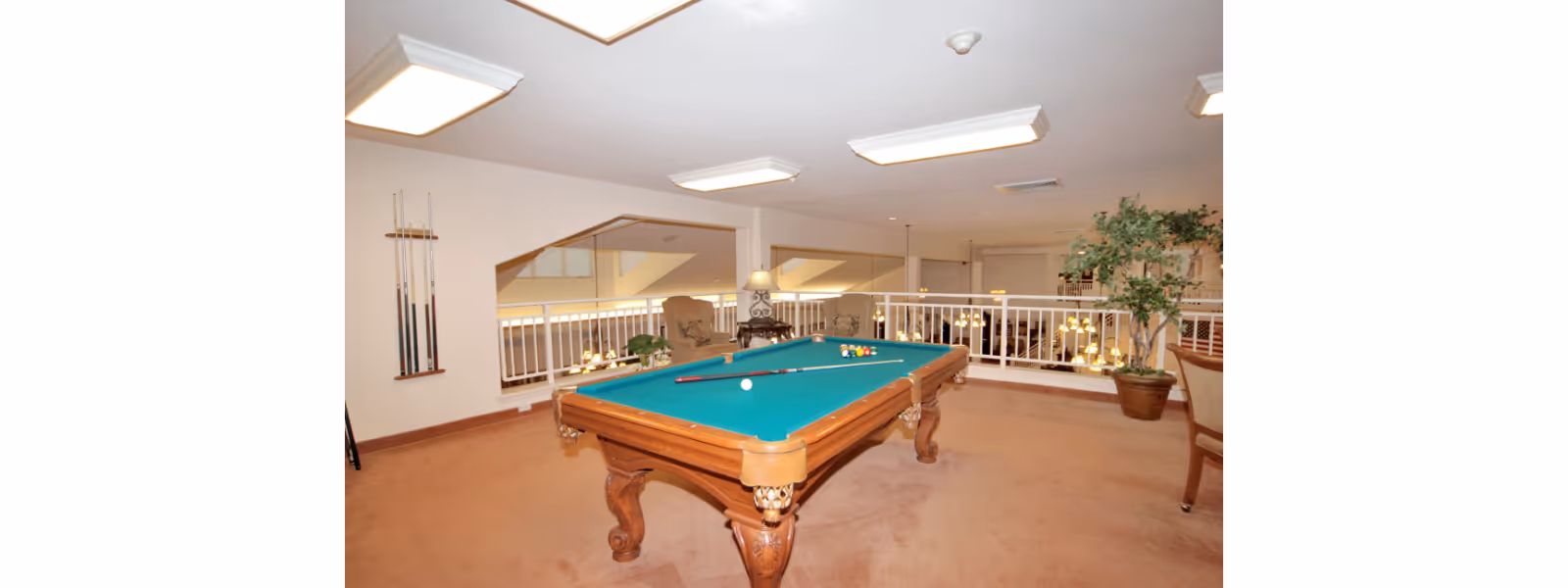 Interior view of a recreational room with a pool table in the center. The pool table has a green felt surface with pool balls arranged and two cues resting on it. The room has beige carpet flooring, white walls, and ceiling lights. There are two armchairs and a small table with a lamp near a white railing overlooking a lower level. A potted plant is placed near the railing on the right side.