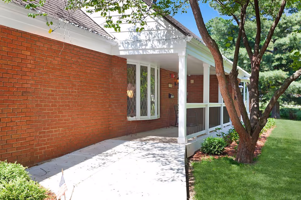 Front exterior of a brick building with a covered white porch, concrete walkway, and surrounding trees and lawn.