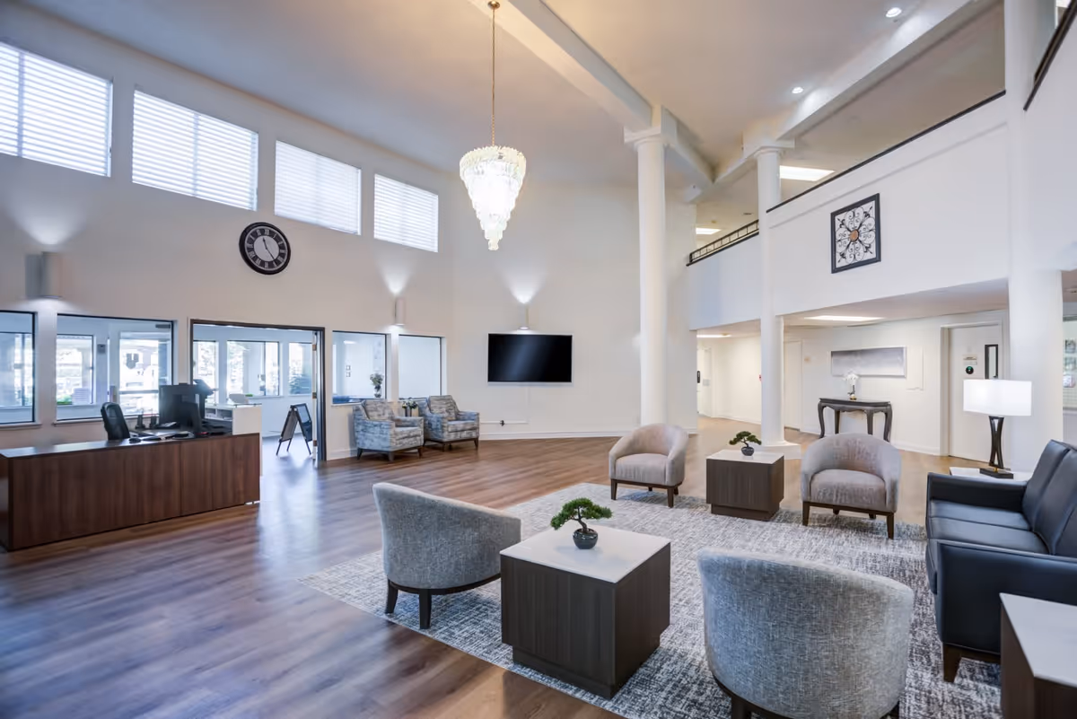 Spacious and well-lit senior living facility lobby with high ceilings, a large chandelier, multiple seating areas including armchairs and a sofa arranged around small tables with plants, a reception desk, a wall-mounted TV, and large windows allowing natural light.