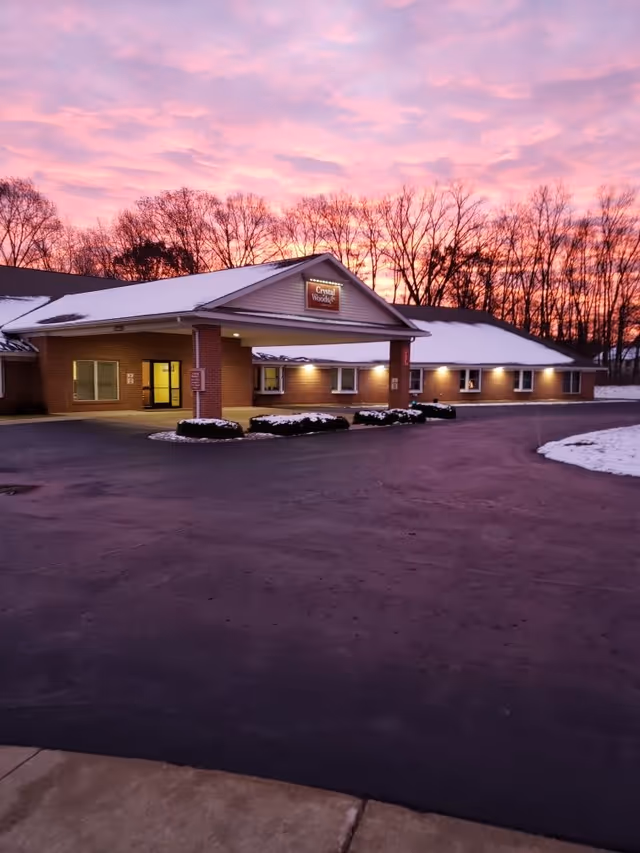 Exterior view of a single-story brick building with a covered entrance and snow on the roof and ground, set against a colorful sunset sky with bare trees in the background.