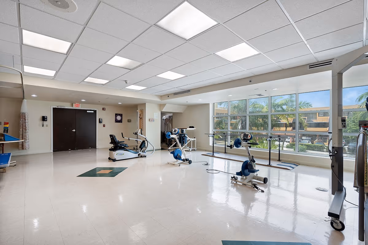 A spacious rehabilitation or physical therapy room with exercise equipment including stationary bikes and parallel bars. Large windows on one side provide natural light and a view of palm trees and a building outside. The room has a tiled floor, white ceiling with fluorescent lights, and a double door at the back.
