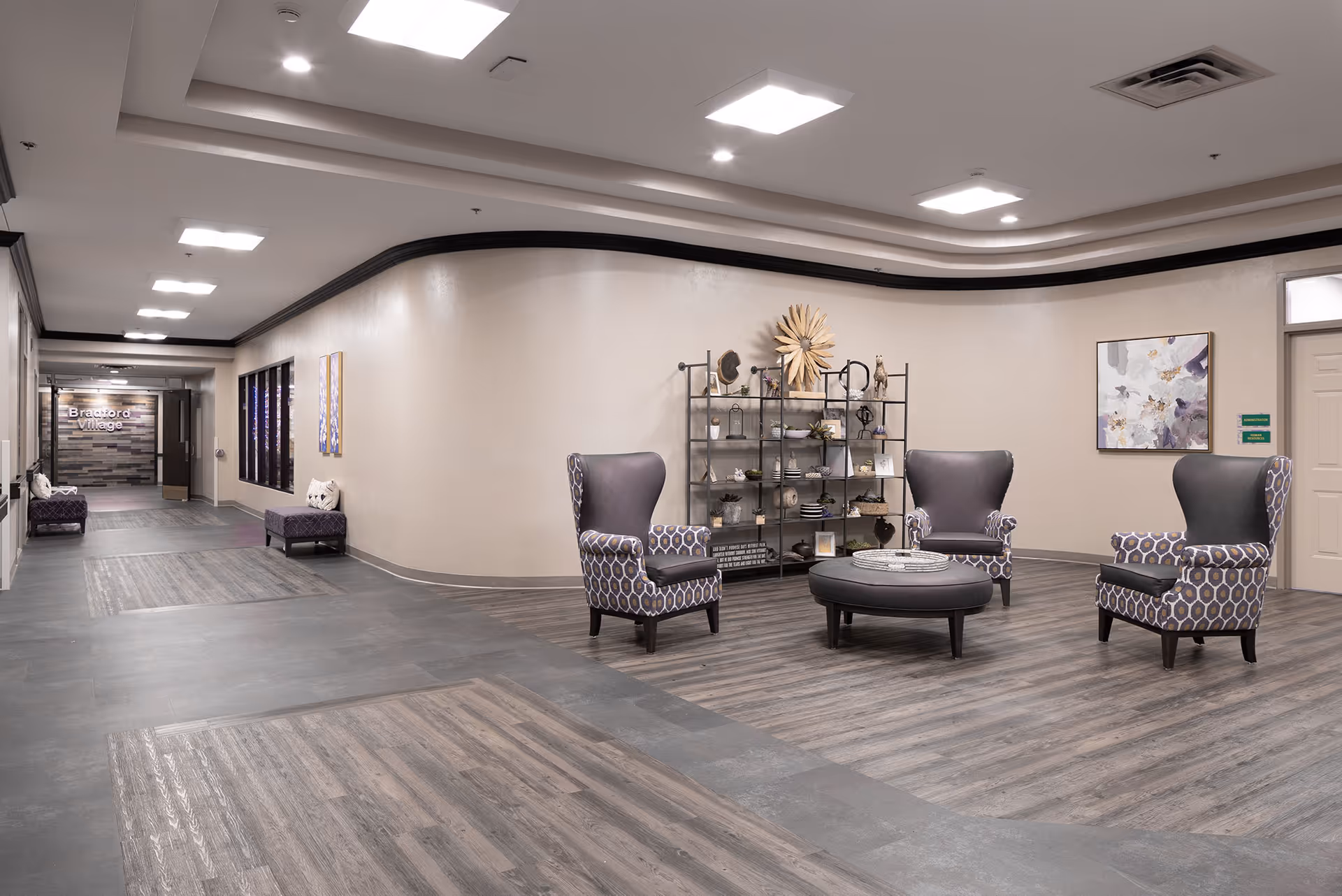 A spacious, well-lit common area in Bradford Village Healthcare Center featuring four patterned armchairs arranged around a round ottoman. Behind the chairs is a decorative metal shelving unit with various ornaments and plants. The walls are light-colored with framed artwork, and the floor has a wood-like finish. In the background, a hallway leads to another area with a sign that reads 'Bradford Village'.