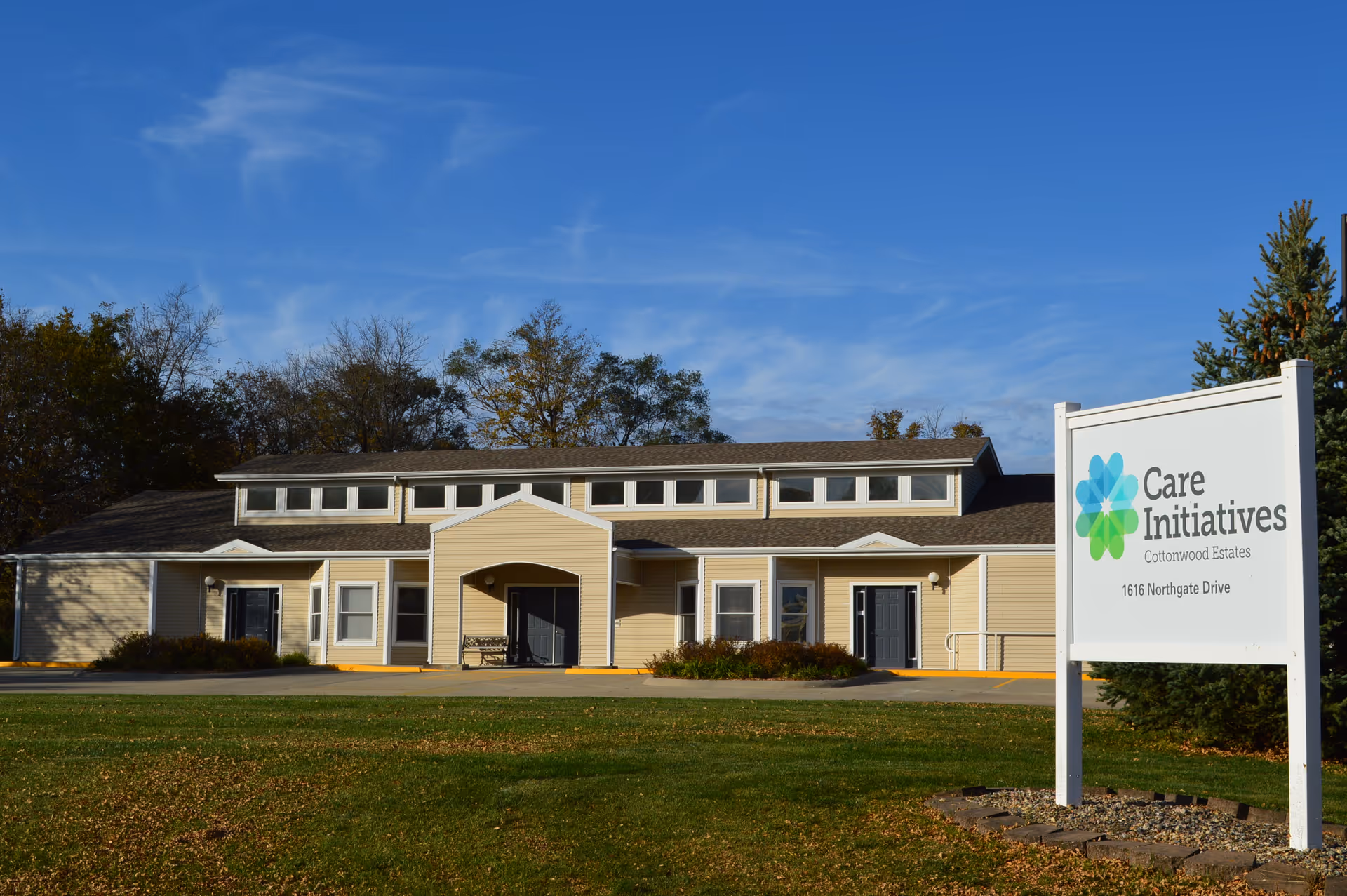 Exterior view of a single-story beige building with multiple windows and two dark-colored doors, surrounded by trees and a grassy lawn under a blue sky. A white sign in the foreground reads 'Care Initiatives Cottonwood Estates 1616 Northgate Drive.'