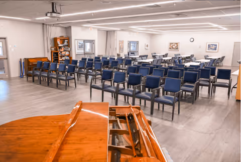 A spacious room with rows of blue chairs arranged facing a wooden piano in the foreground. The room has light-colored walls, framed pictures, a clock on the wall, and a bookshelf in the corner. The ceiling has recessed lighting and a projector mounted.