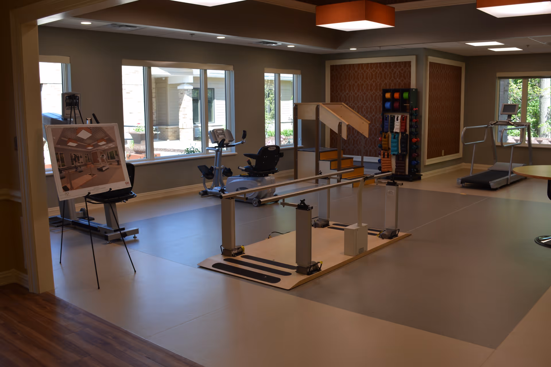 A spacious rehabilitation or physical therapy room with exercise equipment including a recumbent bike, treadmill, parallel bars for walking practice, and a small set of stairs. Large windows allow natural light to enter the room, and a display board with images is set up on an easel near the entrance.