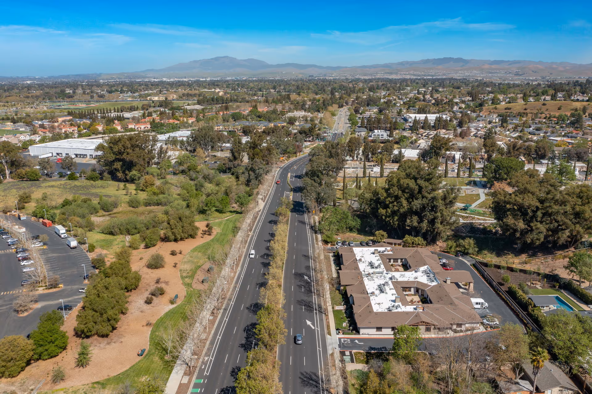 Aerial view of a suburban road with trees, open green spaces, and a memory care facility building under a clear blue sky.