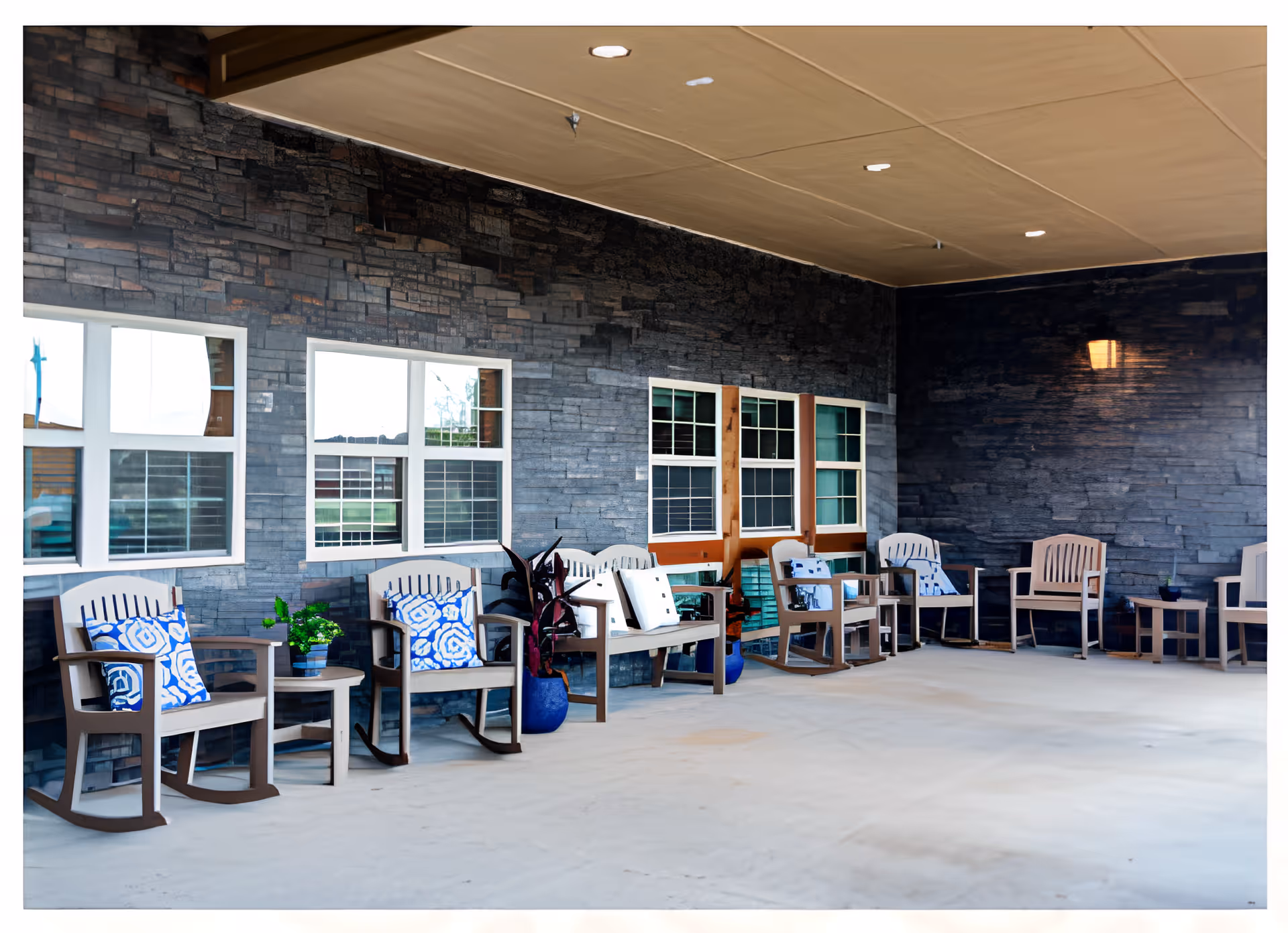 Covered outdoor patio with a row of rocking chairs and small tables along a stone wall under windows.