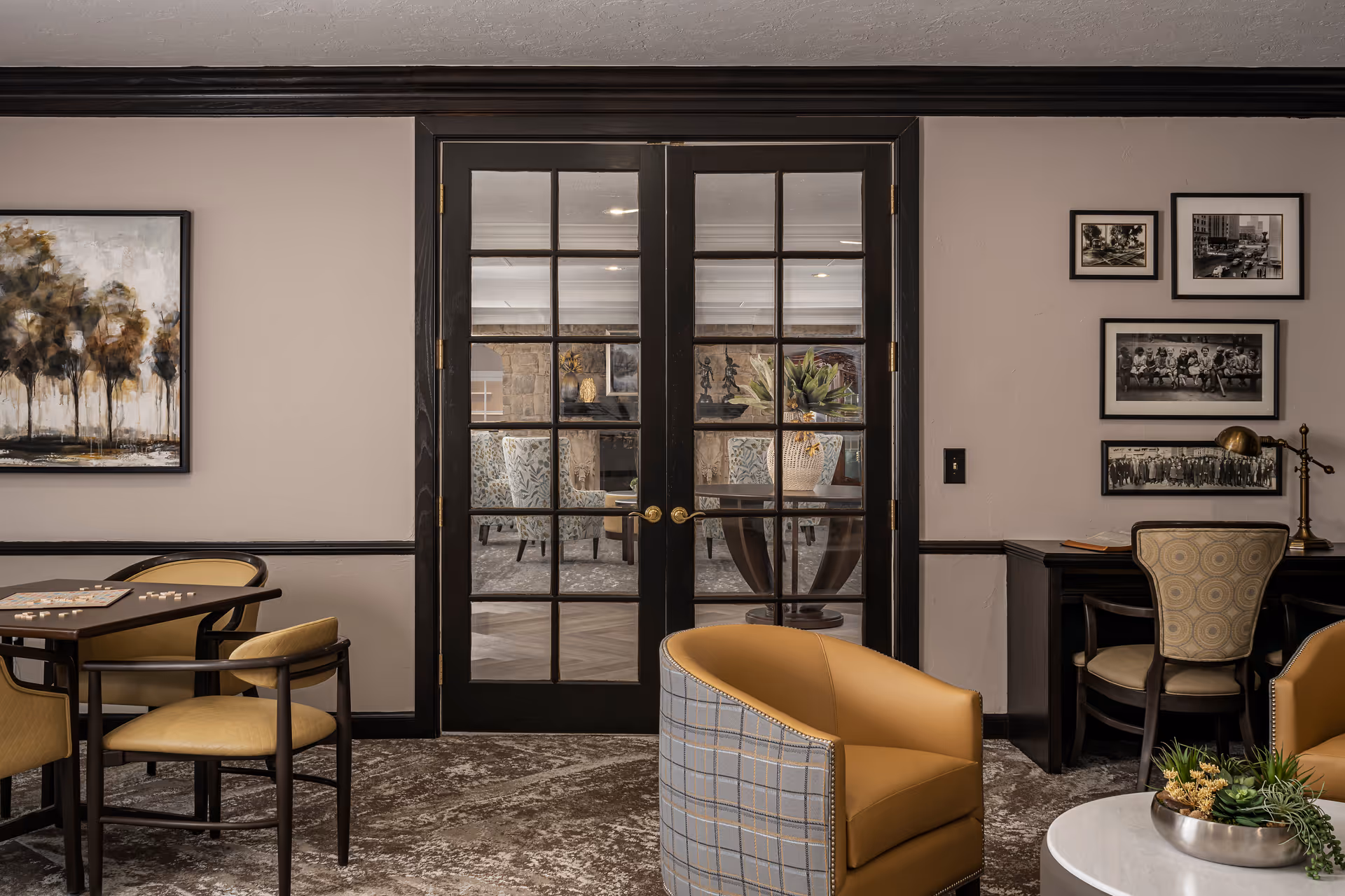 A cozy interior room with a seating area featuring a round table with a plant centerpiece, two armchairs with plaid backs and tan cushions, and a small table with chairs on the left side. The room has a carpeted floor and light-colored walls adorned with framed artwork and black-and-white photographs. In the center, there are black-framed glass double doors leading to another room with additional seating and decor.